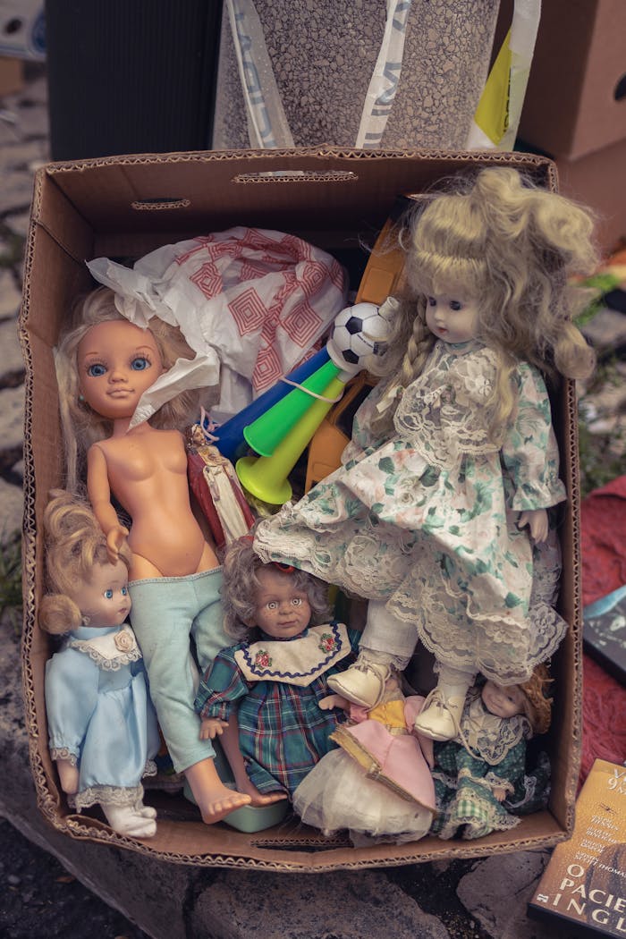 our-story A collection of vintage dolls and toys in a cardboard box placed outdoors.